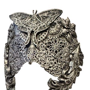 May include: Silver-colored headpiece with intricate filigree and floral designs. A large moth is centered on the crown. The headpiece has a detailed, ornate appearance, with a vintage or fantasy aesthetic.