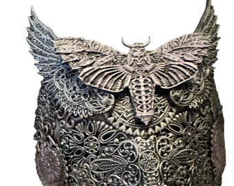 May include: A silver-colored, ornate mask with a large moth or butterfly design at the center. The mask features intricate filigree patterns and detailed wings, creating a gothic or steampunk aesthetic. The mask is likely made of metal.