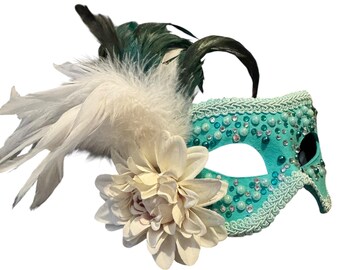 May include: A teal masquerade mask with white and teal embellishments. The mask features a large cream-colored flower and is adorned with white feathers and dark green accents. The mask is decorated with small, round, pearl-like beads.