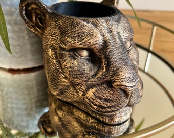 May include: A gold-colored ceramic planter shaped like a jaguar's head. The planter has a black interior and is sitting on a gold-colored tray.