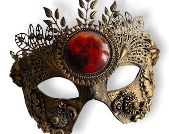 May include: Ornate antique gold masquerade mask with a central red gemstone. The mask features intricate filigree and floral designs, with a crown-like embellishment at the top. The mask is designed to cover the eyes and nose.