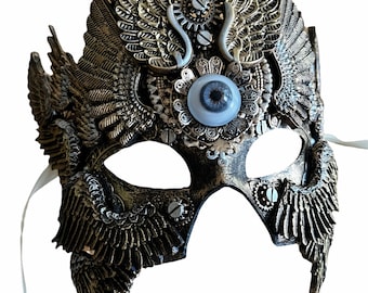 May include: A gold and black masquerade mask with intricate details, including wings, gears, and a large eye in the center.