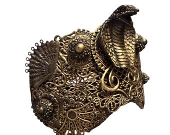 May include: Ornate, antique gold-colored cuff bracelet featuring a detailed cobra design. The cuff has intricate filigree and floral patterns, with a fan-shaped element on the side. The cobra head is raised, adding dimension.