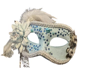 May include: A light blue masquerade mask with white and silver accents. The mask is decorated with blue and pearl-like beads, a white flower, and white feathers. The edges are trimmed with silver braid.