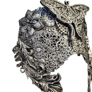 May include: A silver-toned, ornate headpiece with a butterfly and floral design. The headpiece features intricate detailing, including a butterfly at the top, floral accents, and delicate filigree patterns. The overall design is elaborate and detailed.