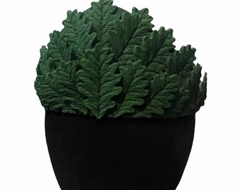 May include: A dark green leaf crown, resembling a laurel wreath, sits atop a black form. The crown is made of detailed, textured leaves, creating a naturalistic appearance. The leaves are a deep, forest green color, and the crown is likely a costume accessory.