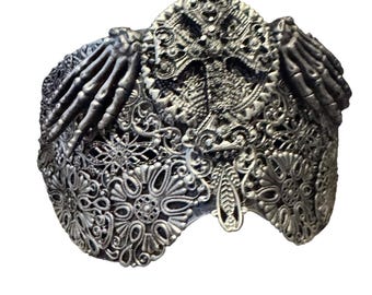May include: Silver-toned, ornate headpiece with a gothic aesthetic. The headpiece features intricate filigree patterns, a central cross design, and skeletal hand accents on each side. The overall design is detailed and textured.