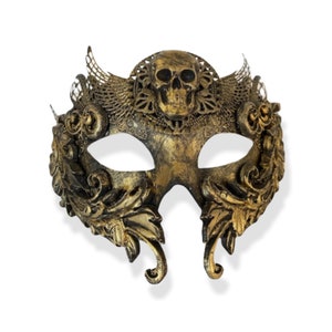 Op de afbeelding: Een goudkleurig masker voor een gemaskerd bal met een schedel in het midden bovenaan. Het masker heeft sierlijke, wervelende ontwerpen en veerachtige accenten. De ooggaten zijn ovaal. Het masker is waarschijnlijk gemaakt van een hard materiaal.