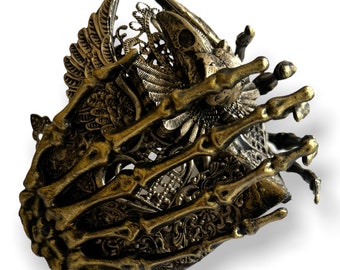 May include: A gold-toned metal bracelet with a skeletal hand design. The bracelet features intricate details, including a bird skull and wings.