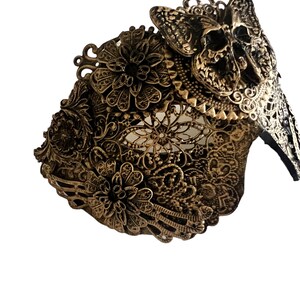 Puede incluir: Una m&aacute;scara de metal de color dorado con un dise&ntilde;o de calavera y mariposa. La m&aacute;scara est&aacute; intrincadamente detallada con patrones florales y tiene una apariencia delicada y de encaje.