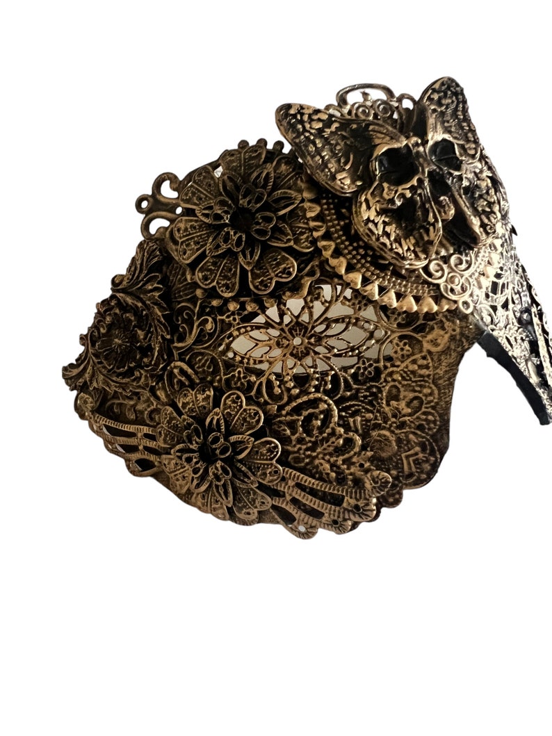 Puede incluir: Una m&aacute;scara de metal de color dorado con un dise&ntilde;o de calavera y mariposa. La m&aacute;scara est&aacute; intrincadamente detallada con patrones florales y tiene una apariencia delicada y de encaje.