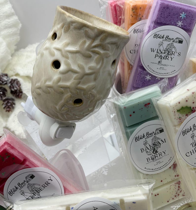 May include: A white ceramic wax warmer with a leaf design and a white plug-in cord. The warmer is surrounded by various scented wax melts in clear packaging. The wax melts are labeled with the names "Winter's Pantry", "Balm & Berry", and "Cranberry".