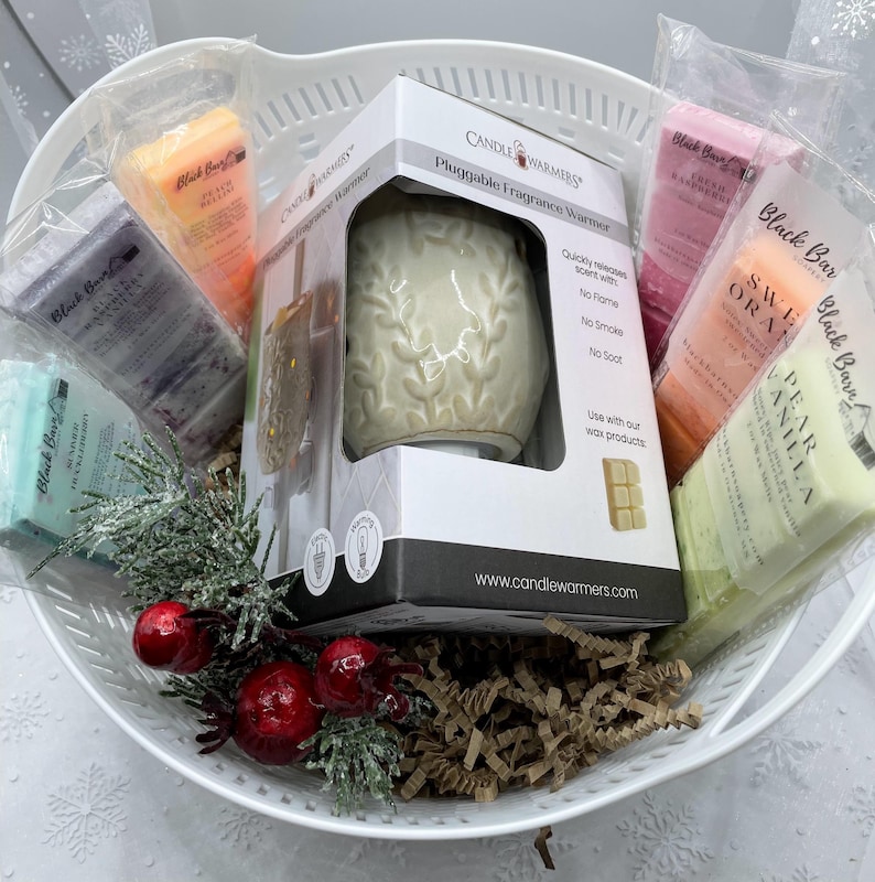 May include: A white ceramic plug-in fragrance warmer with a floral design, surrounded by a variety of scented wax melts in a white wicker basket. The wax melts are labeled "Black Bar" and include scents like "Sweet Orange" and "Pear Vanilla".