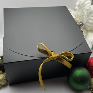 May include: A black gift box with a gold ribbon tied in a bow. The box is sitting on a white surface with green and red ornaments.