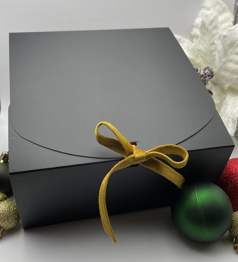 May include: A black gift box with a gold ribbon tied in a bow. The box is sitting on a white surface with green and red ornaments.