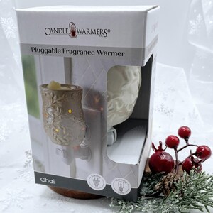 May include: A white ceramic plug-in fragrance warmer with a floral design. The warmer is labeled "Chai" and has a light bulb icon. The warmer is on a wooden surface with a red berry sprig.