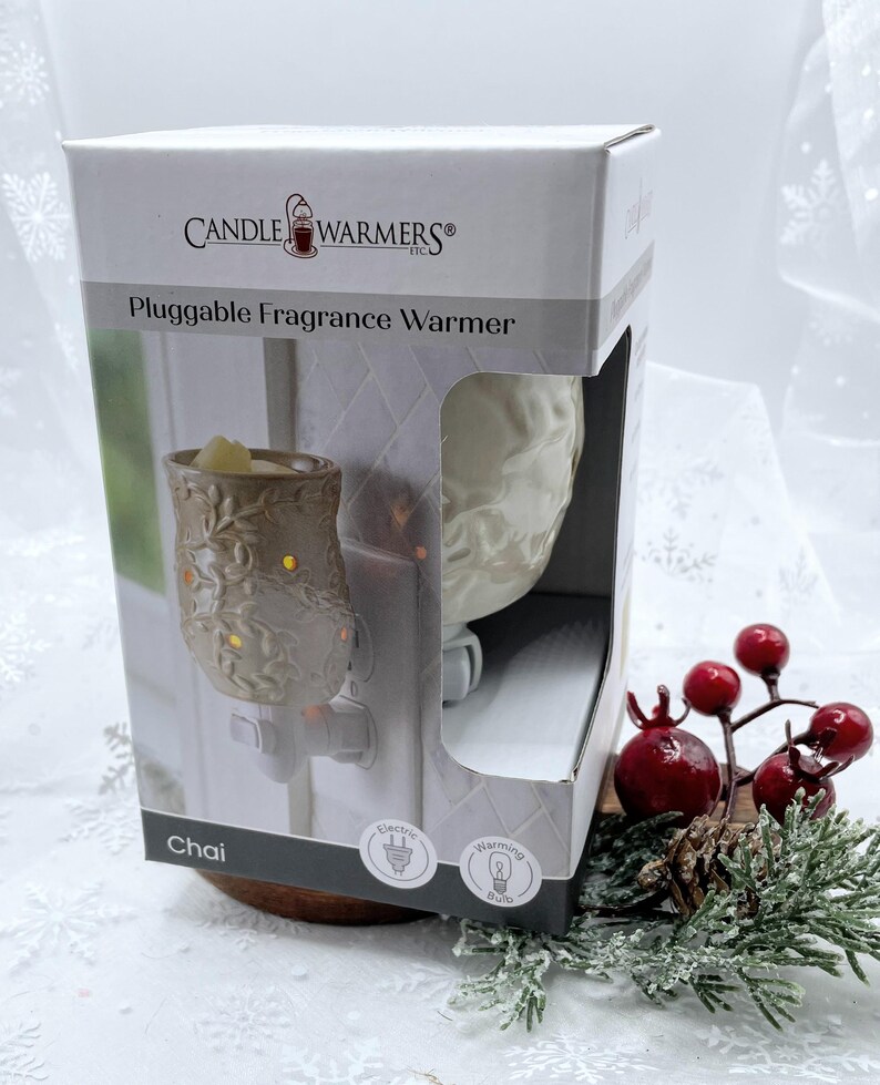 May include: A white ceramic plug-in fragrance warmer with a floral design. The warmer is labeled "Chai" and has a light bulb icon. The warmer is on a wooden surface with a red berry sprig.