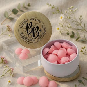 May include: A white metal tin filled with small, pink, dome-shaped wax melts, accompanied by a round wooden lid featuring a black logo with the text Wild Rose - Dew Drops and Black Barn Soapery, surrounded by small white flowers and green leaves.
