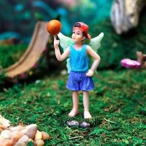 May include: A small fairy figurine with wings, wearing a blue and white dress, red hat, and holding an orange ball. The fairy is standing on a green mossy surface.