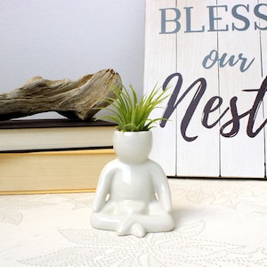 May include: White ceramic planter shaped like a person sitting in a meditative pose. The planter has a small opening at the top for a plant. A small air plant is growing in the planter. The planter is sitting on a white surface with a wooden sign that says "Bless our Nest" in the background.