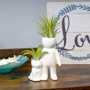 May include: White ceramic planter shaped like a person holding a white ceramic planter shaped like a cat. Both planters are filled with green air plants.