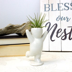 May include: White ceramic planter shaped like a person with arms raised, holding a small air plant. The planter is standing on a white base. The background includes a wooden sign with the text "Bless our Nest".
