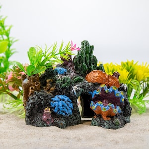 May include: A dark gray and brown resin aquarium ornament shaped like a cave with a blue and green clam shell, a brown and orange shell, and green and blue coral. The ornament is surrounded by green plastic plants.