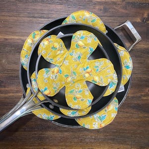 May include: A stack of three black frying pans with silver handles, each containing a yellow, flower-shaped pot holder. The pot holders feature a pattern of lemons, green leaves, and white flowers. The pans are on a dark wooden surface.
