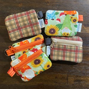 May include: Four small fabric zipper pouches featuring two distinct patterns: a tan, brown, and red plaid design and a light green fabric printed with sunflowers, mushrooms, and gnomes. Each pouch includes a small side tab and a metal zipper closure.