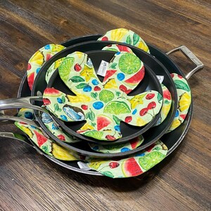 May include: A set of black frying pans with silver handles, each containing a colorful, fruit-patterned fabric liner. The liners feature illustrations of watermelon, cherries, blueberries, and other fruits on a light green background.