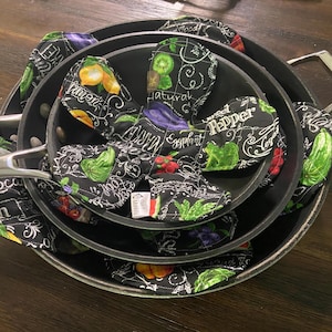 May include: A set of nested black metal pans featuring protective fabric pan protectors. The protectors are black with a white grid pattern and colorful illustrations of fruits and vegetables, including lemons, eggplants, and peppers with text labels.