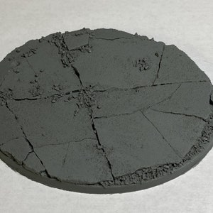 May include: A round, gray, textured, cracked, and broken base for miniature figures. The base is made of a hard plastic material and has a rough, uneven surface.
