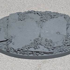 May include: A gray oval-shaped plastic base with a textured surface that resembles cracked pavement. The base is designed for miniature figures and games.