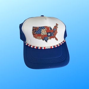 May include: A blue and white trucker hat with a patriotic design. The hat features a white front panel with a patchwork map of the United States. The brim is blue, and the hat has a mesh back. The word "USA" is spelled out on the brim.