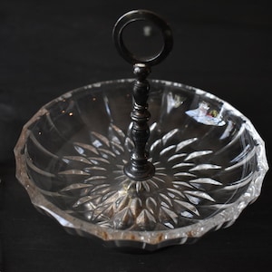 May include: A clear, cut-glass serving dish with a dark metal handle. The dish has a starburst pattern on the base and a scalloped edge. The handle is attached to the center of the dish.