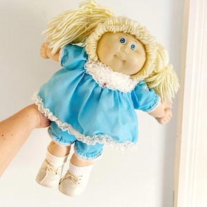 May include: A vintage doll with a soft, round face, blue eyes, and blonde yarn hair. The doll wears a light blue dress and matching shorts trimmed with white lace. It has white socks and cream-colored shoes.