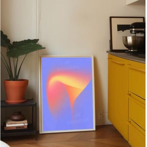 May include: A framed art print featuring a gradient design in shades of orange, yellow, and pink against a blue background. The text "JD DESIGNS" is visible on the print. The artwork is displayed in a room with a plant, a black side table, and yellow cabinets.
