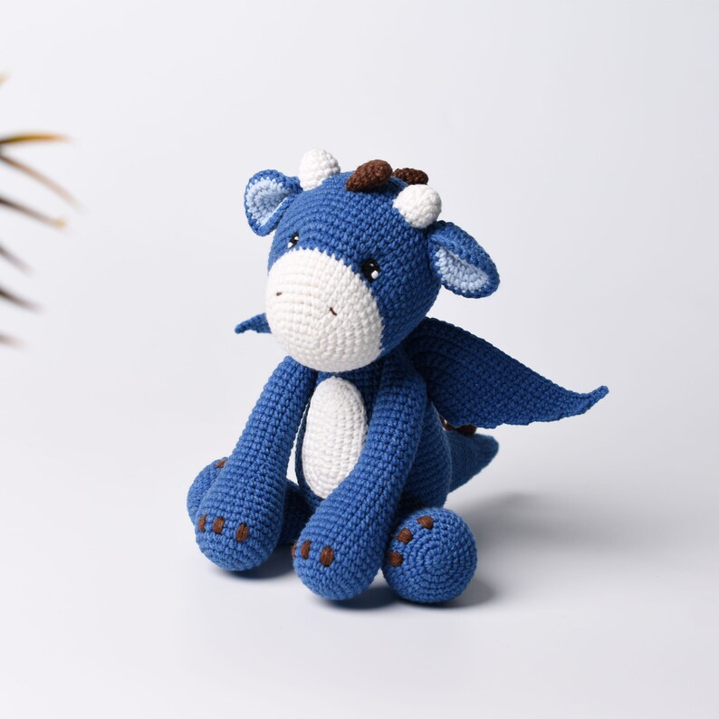 May include: A blue crocheted dragon toy with white accents and brown eyes. The dragon is sitting with its wings spread out.