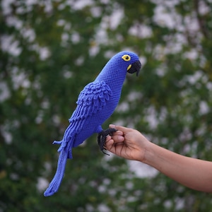 May include: A handmade, royal blue crochet parrot with a black beak and yellow eye accents. The bird's wings and tail feathers are detailed. The parrot is held in a hand against a blurred green background.