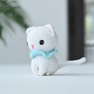 May include: White crocheted cat with a light blue scarf. The cat has black eyes and a pink nose.