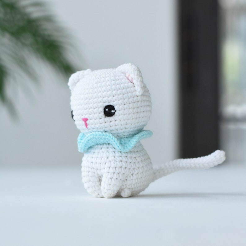 May include: White crocheted cat with a light blue scarf. The cat has black eyes and a pink nose.