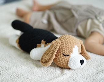 May include: A crocheted beagle dog plush toy with brown, black, and white fur, lying on a white fuzzy blanket. The dog has its eyes closed and appears to be sleeping.