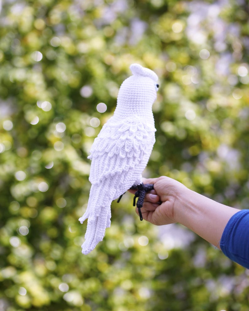 以下が含まれることがあります： 手作りの白いかぎ針編みの鳥で、羽のディテールと小さなとさかがあります。鳥は手に止まっており、その複雑なデザインを示しています。鳥の足は濃い灰色です。背景は柔らかくぼやけた緑色です。