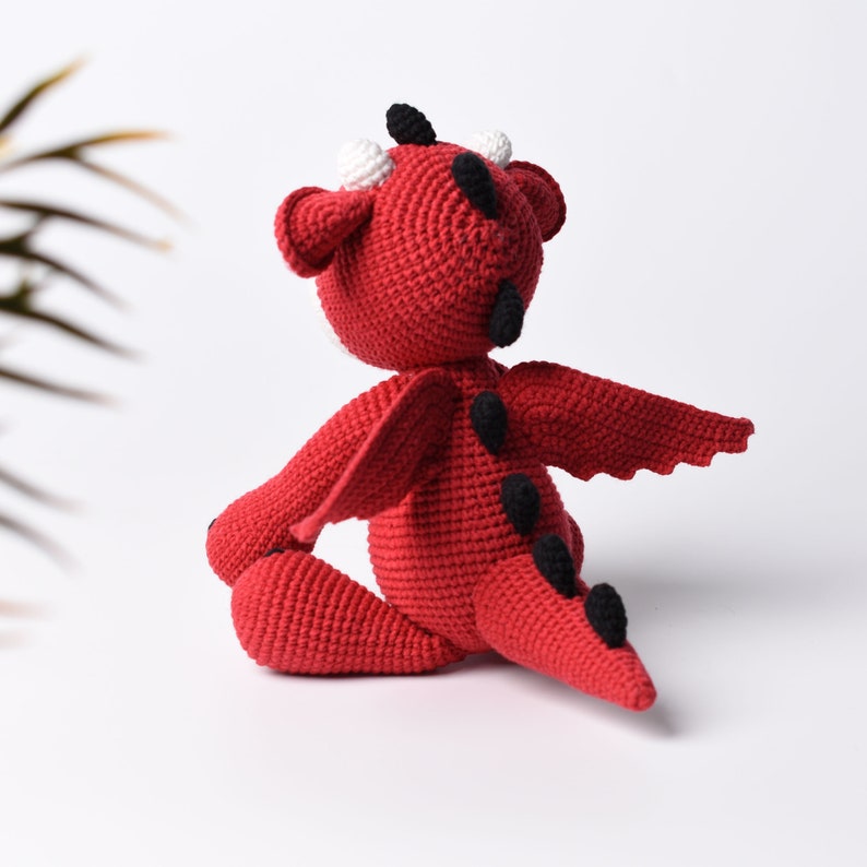 May include: A red crocheted dragon toy with black eyes, white horns, and a black tail. The dragon has wings and is sitting on a white surface.