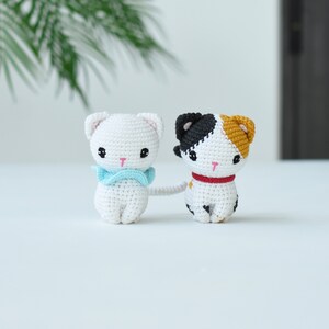 May include: Two crocheted cat toys, one white with a blue bow and the other white with black and orange patches. The cats are sitting on a white surface.