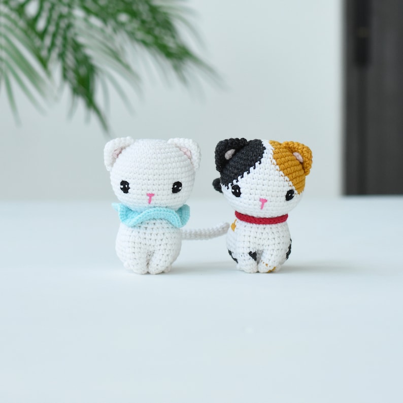 May include: Two crocheted cat toys, one white with a blue bow and the other white with black and orange patches. The cats are sitting on a white surface.