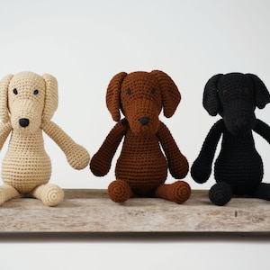 May include: Three crocheted dog toys, one beige, one brown, and one black, sitting on a wooden surface.