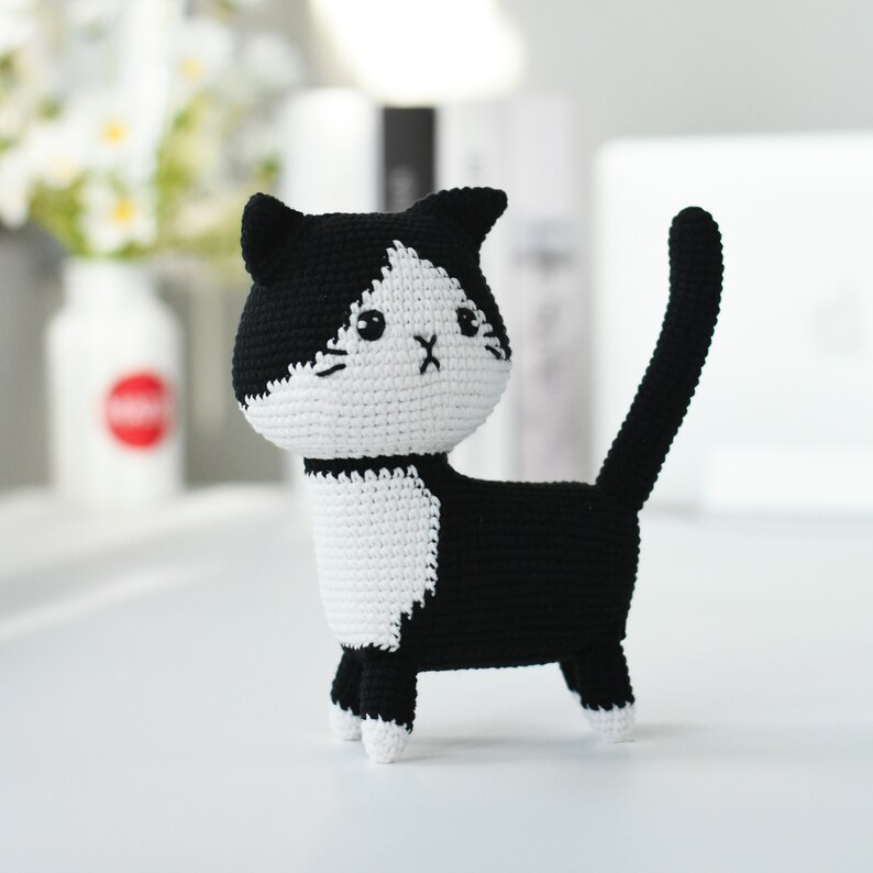 May include: A black and white crocheted cat toy with a sad expression. The cat is standing on a white surface.