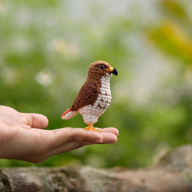 May include: A small, handcrafted crochet bird figurine featuring a brown back, a white chest with brown speckles, a black beak, and small yellow feet. The bird is depicted in a standing position, showcasing detailed yarn stitching throughout its body.
