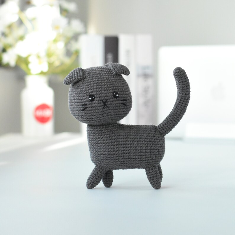 May include: A gray crocheted cat toy with black eyes and a stitched-on nose. The cat is standing on a light blue surface.